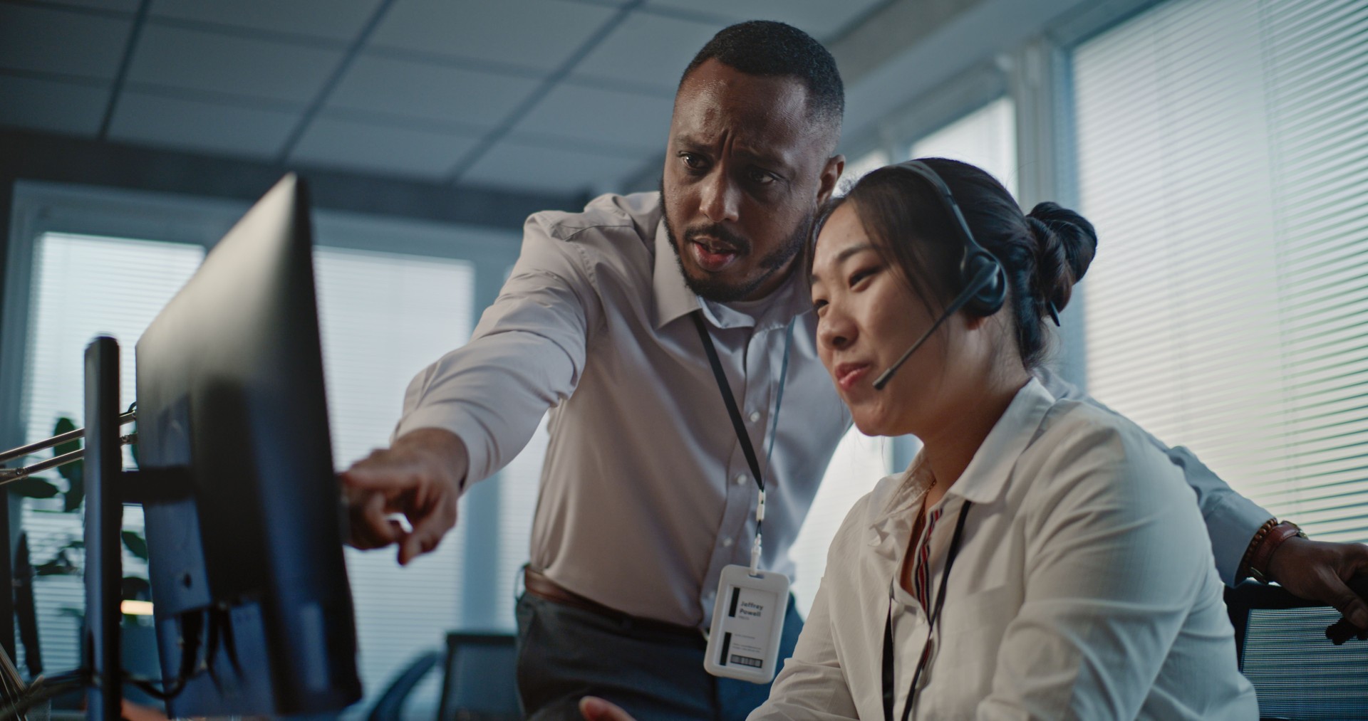 Asian helpdesk specialist in headset uses computer, talks to African American colleague
